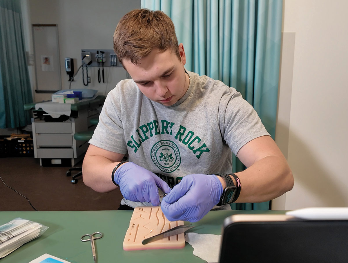 A student in a Slippery Rock University t-shirt and purple medical gloves practices suturing on a synthetic skin training pad in a medical exam room.