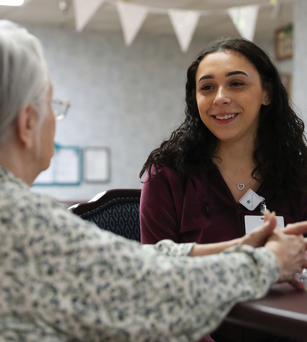 A smiling young woman wearing an ID badge holds hands across a table with an elderly woman.